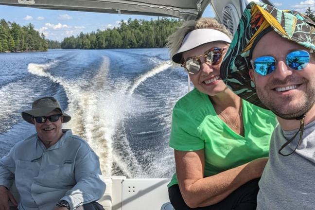 Three people smiling on a boat ride with a lake and trees in the background.
