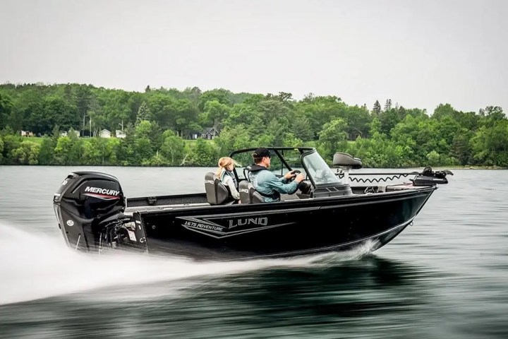 Black boat with two people speeding on a lake against a backdrop of trees.