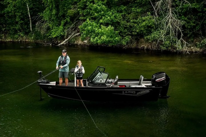 Two people fishing on a boat in a green, forested area with clear water.